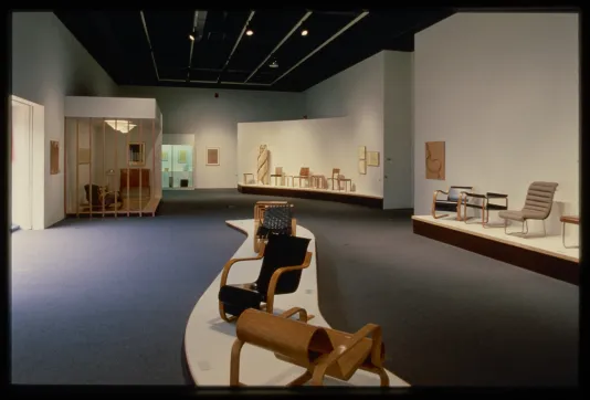 A wave shaped pedestal displays wooden chairs in the center of the gallery. The walls are lined with more chair designs.