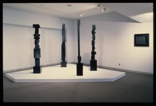 Sculptural towers made from wood painted black sit on a pedestal. A small black wood sculpture hangs on the opposite wall. 