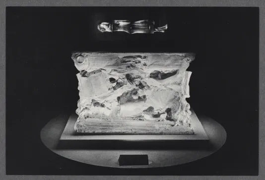 A black and white photo of a ceramic sculpture. The sculpture is a cube shape with faded details, spotlit on a pedestal.