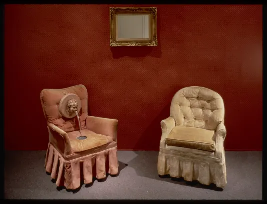 Two decorative chairs set in front of a red wall with a gold framed mirror hung between them.