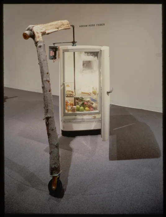 A tree branch on wheels is attached to an open fridge containing various food products.