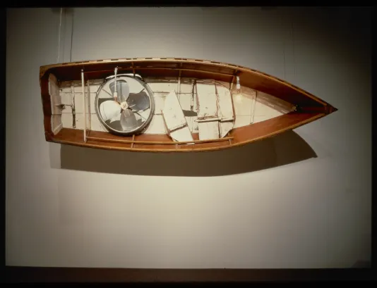A wooden toy boat is hung sideways on the wall with a fan or propeller rests inside.