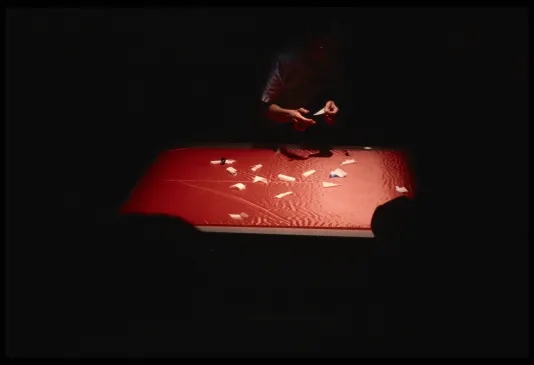A performer places triangular pieces of paper on a spotlit red table top. Viewers sit in front in the darkness.