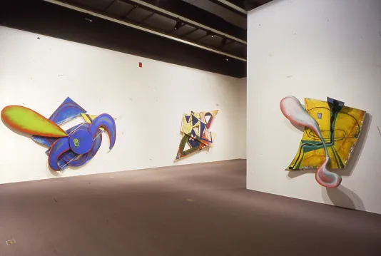 Three bright colored irregular shaped abstract paintings hung on walls, part of one canvas extends outward casting a shadow.