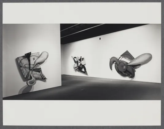 A black and white photo of three unique shaped abstract paintings hung on walls, part of one extends outward casting a shadow.
