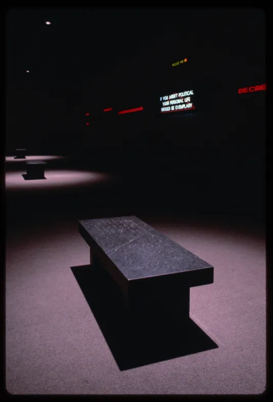 Sign reading “If you aren’t political your personal life should be exemplary” hung in a dark room with three benches spotlit.