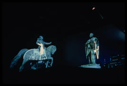 Two pictures of statues overlaid with projections, one of a man on a horse and one of a man standing, hang in a pitch black room.
