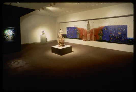 Wooden sculpture sits in middle of room, on walls are lit installation, colorful wall installation, and small sculpture.