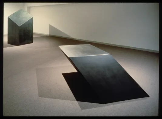Two large, black, metal, geometric sculptures sit in a gallery.