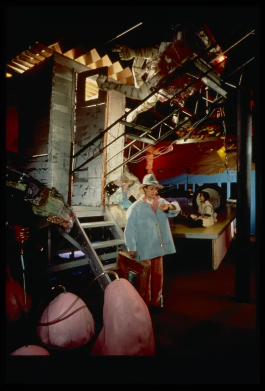 Person in hat and blue coat look at watch while woman sits on a platform in an abstract mechanical landscape installation.