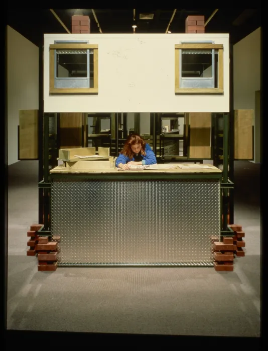 A visitor sits inside a cubicle made from industrial materials while reading a newspaper.