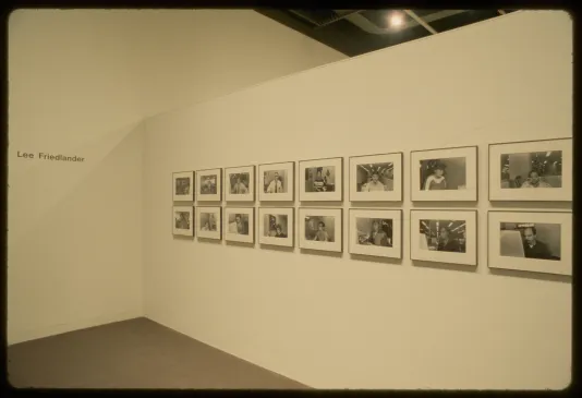 Sixteen small black and white photos of people hang in two rows on a gallery wall. Wall text reads “Lee Friedlander”