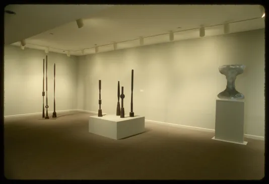 Three groupings of sculptures stand by the gallery wall. Two are tall pillar-like sculptures and one sculpture stands alone. 