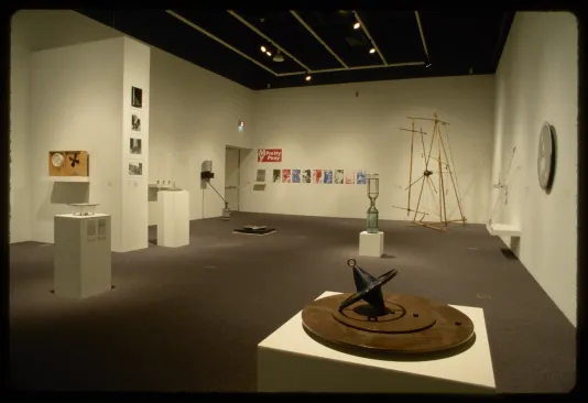 An installation view of hanging works and sculptures. Many sculptures take on the form of clocks, dials, or hourglasses.