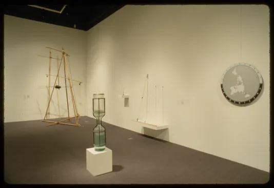 An hourglass sits on a pedestal. A clock is placed on the wall to the right and a geometric sculpture is on the left.