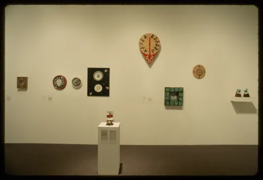 A variety of clocks are hung on the wall at different heights while a small sculpture sits on a pedestal in front.