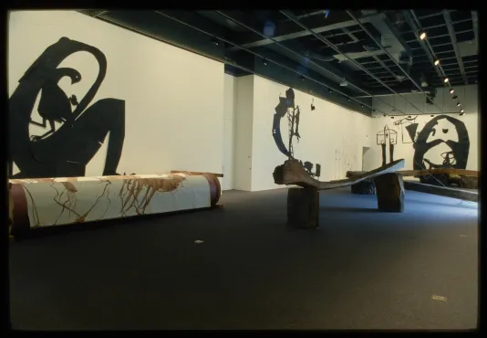 Gallery view of four giant black paper cut outs of abstract figures on the walls, and three wooden slabs and logs on the floor.