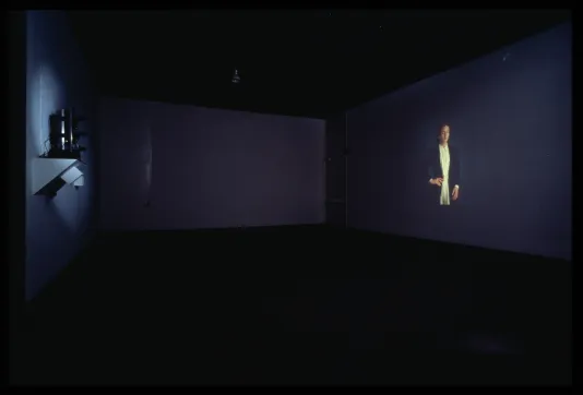 A dark gallery space shows a projected film. In the still there is a figure posing with their hand on their hip.