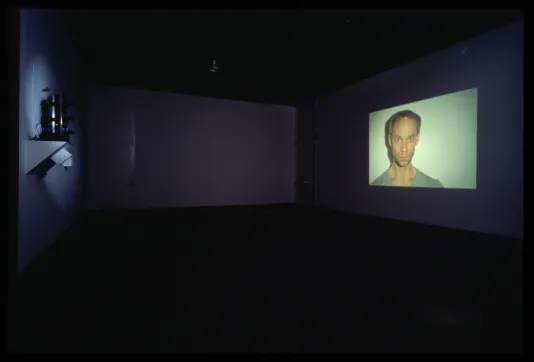 A dark gallery space shows a projected film. In the still is a bust of a figure staring directly at the camera.