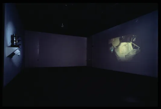 A dark gallery space shows a projected film. In the still is an empty hospital bed with the covers in disarray.