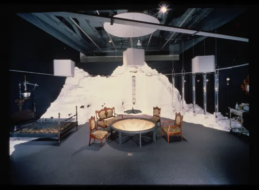 An installation of a room with a table, chairs, and bed. These elements sit before a constructed white mountain.