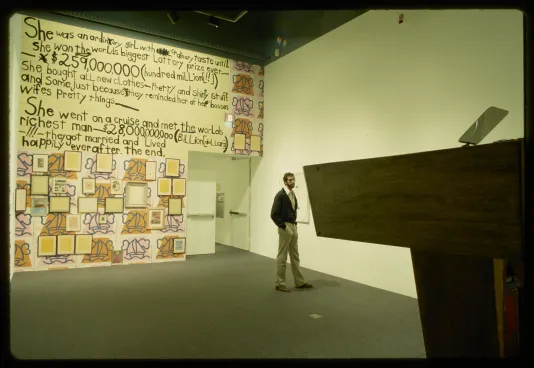 A podium stands in the foreground as a visitor looks on. The entrance wall behind them displays a large hand written letter.