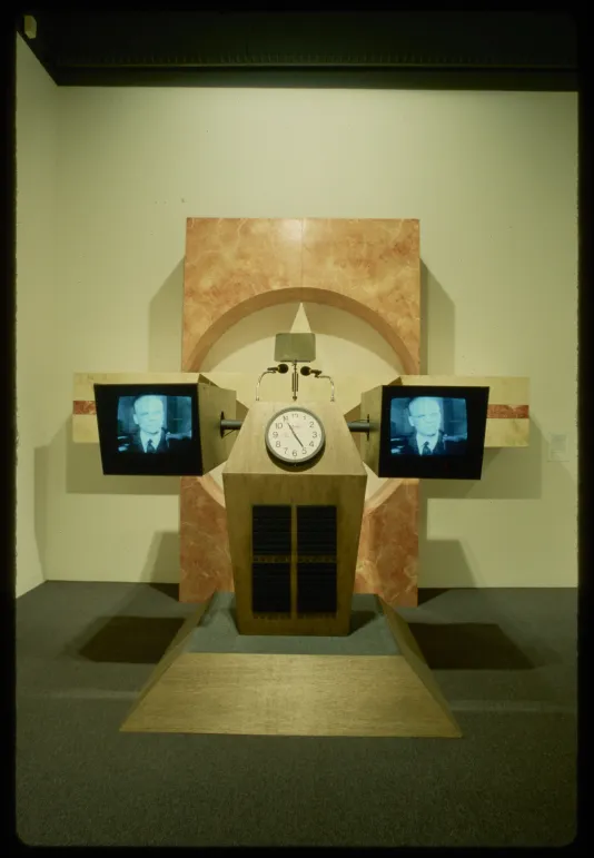 A podium has a clock with two monitors on each side showing a television broadcast.