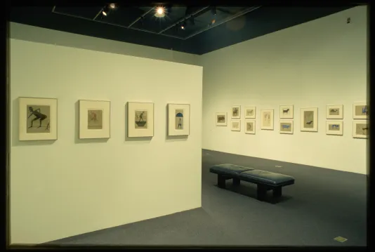 Framed drawings of figures and animals are hung on the gallery walls while a bench is placed for viewing the work. 