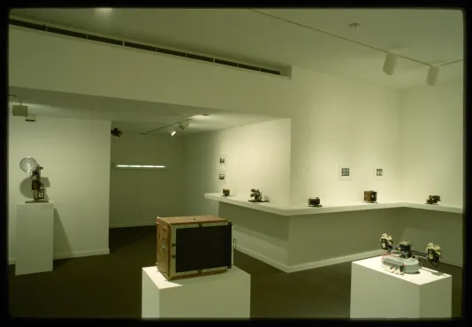 Different types of cameras sit on pedestals and on shelves. Black and white photographs are pinned to the gallery wall.