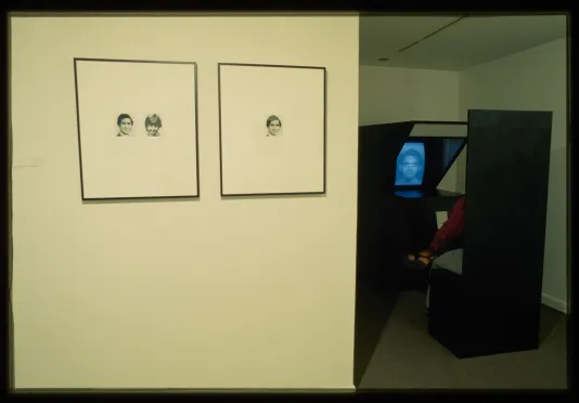 Two images hang on the gallery wall while a photobooth is set up in a dark corner of the gallery. 