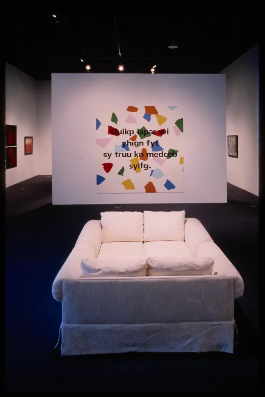 Two white couches face each other in front of a text based work with blotches of different colors.
