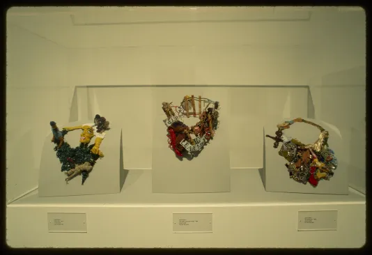 Three necklaces sit on stands within a glass case. Each one is made from different materials with abstract imagery.