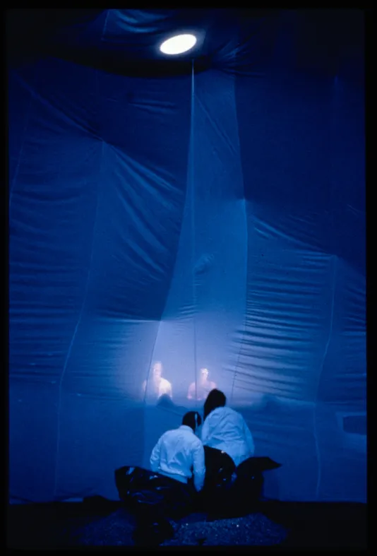Two performers dressed in white emerge from encasings while watching a projection of two people before them.