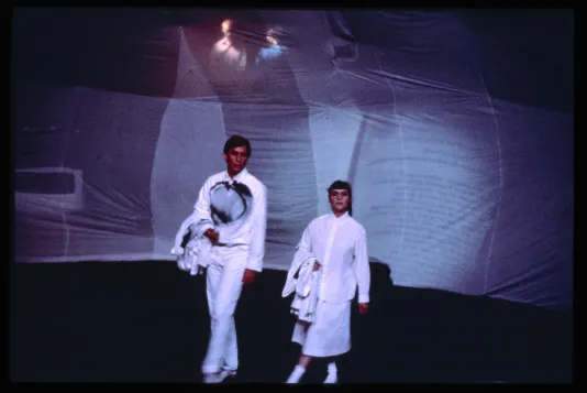 Two performers look at the audience. Behind them are large sheets onto which a projection is played. 