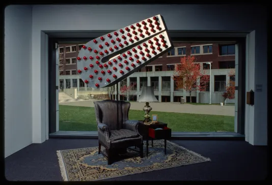 Giant metal magnet with red lightbulbs in midair above leather chair and desk and lamp on rug, in front of large window. 