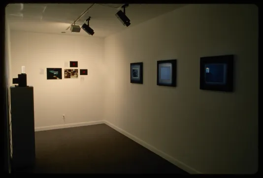 A small monitor stands on a pedestal while other screens are mounted to the gallery wall. Images are hung between them. 