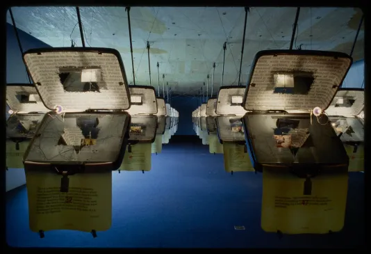 Identical opened suitcases are hung from the ceiling in rows. Each has text projected on the inside. 