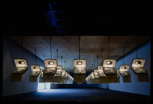 Identical opened suitcases are hung from the ceiling in rows. Each has text projected on the inside.