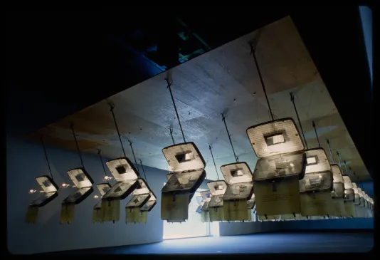 Identical opened suitcases are hung from the ceiling in rows. Each has text projected on the inside.