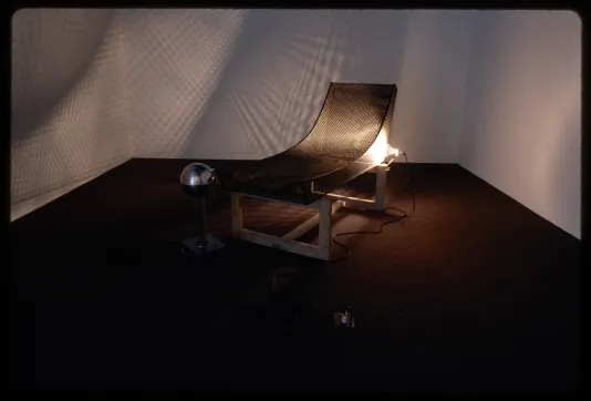 A wire mesh sculpture of a reclined chair on a wooden base. A light shines through, projecting a fence pattern on the wall. 