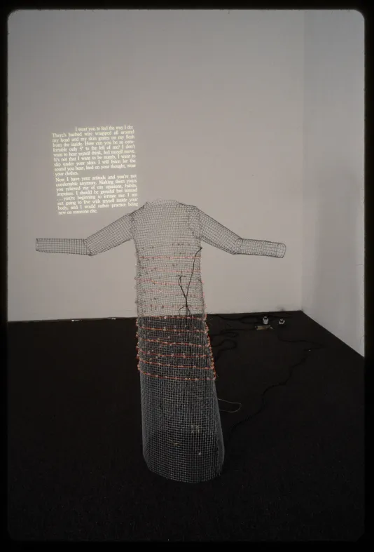 A wire mesh sculpture made to look like a standing body. Projected text is lit on the wall behind the sculpture.