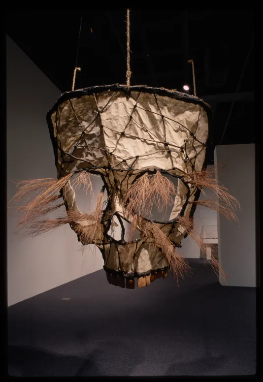 The front of a skull is sculpted from twigs and paper hanging from the ceiling.  From the eyes sprouts dry red grass. 