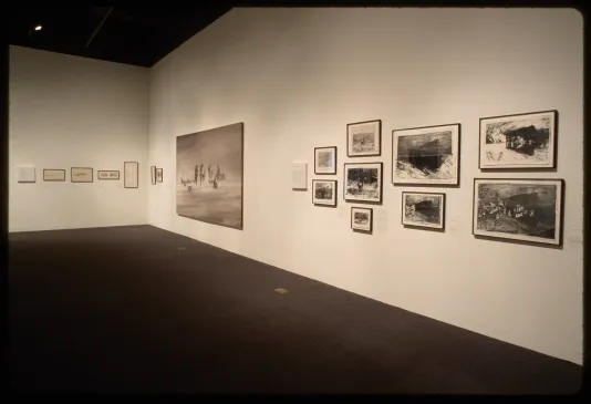 Framed text hangs adjacent to large monochrome oil painting of nomads in a desert and nine framed black and white photographs. 