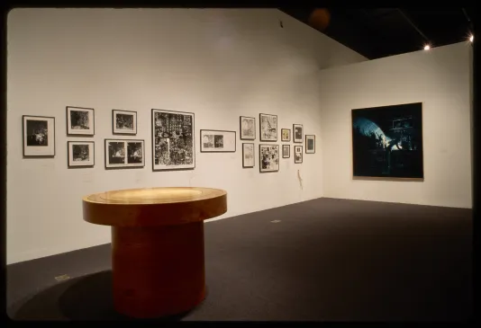 Circular wooden table sits gallery with sixteen framed black and white photographs and large square photograph on walls. 