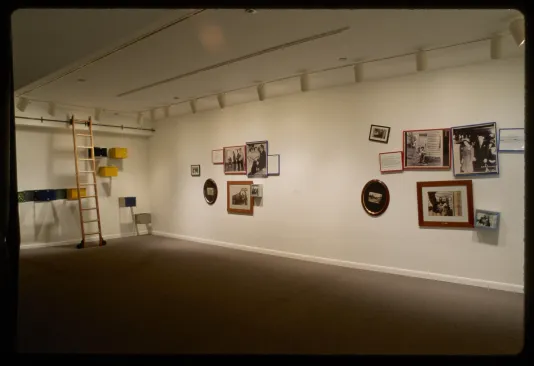 Two groups of eight black and white photos hang on adjacent gallery wall to wooden ladder in front on colorful boxes on wall.