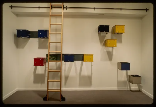 Wooden ladder stands in front of blue, yellow, red, and green metal boxes hung in asymmetric groups on the gallery wall. 