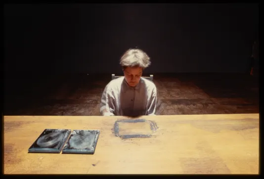 Person with short hair sits at table staring at rectangle outline made of dust, with two stacks of mirrors to their right.
