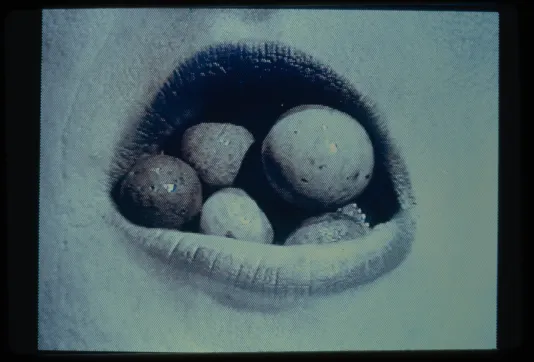 Blue-tinted video projection of close up of person’s open mouth showing five small spherical stones of various shades inside.