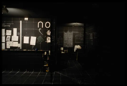 Black room with chalk drawings of furniture to the right, and diagrams and papers hung on the wall to the left. White chalk grid lines entire space.
