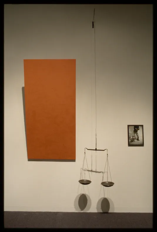 Scales hang from chain in front of wall with large orange rectangle hanging at angle on left and small picture of person on right.